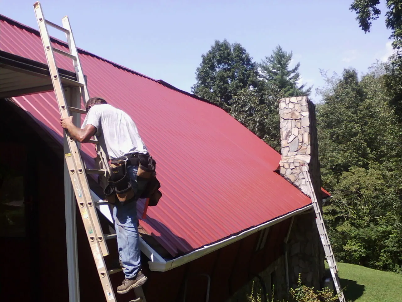 Roofer on a ladder beside a red metal roof and stone chimney — Soffit & Fascia Repair by Roofing Brothers in Ponca City